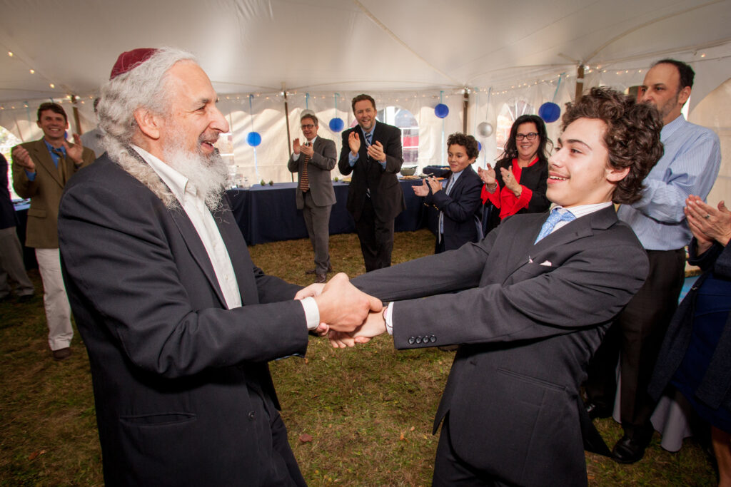 An older man and a teenage boy in suits clasp hands and smile at each other under a tent, surrounded by clapping people.