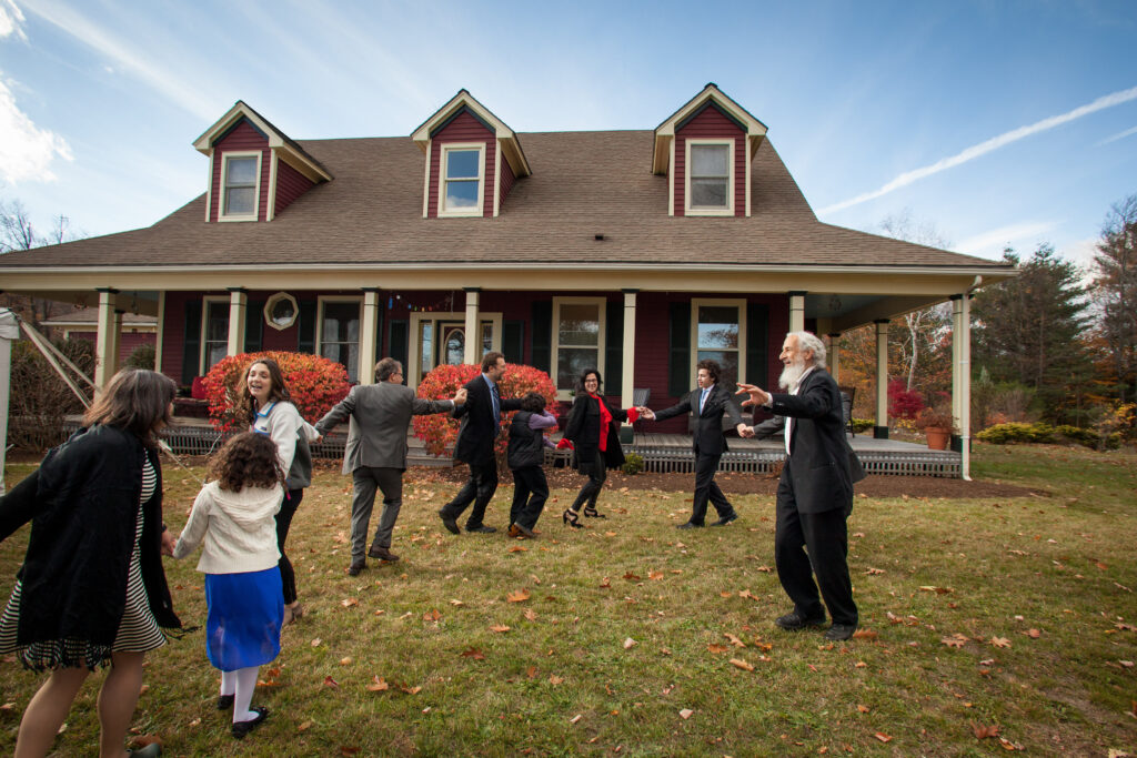 A group of people, both adults and children, stand and interact on the lawn in front of a two-story house on a clear day.