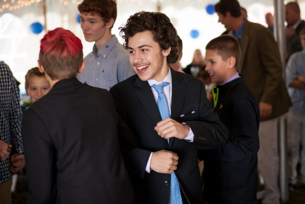 Teenagers dressed in suits laugh and interact at an indoor event, with one boy in focus smiling while adjusting his jacket.
