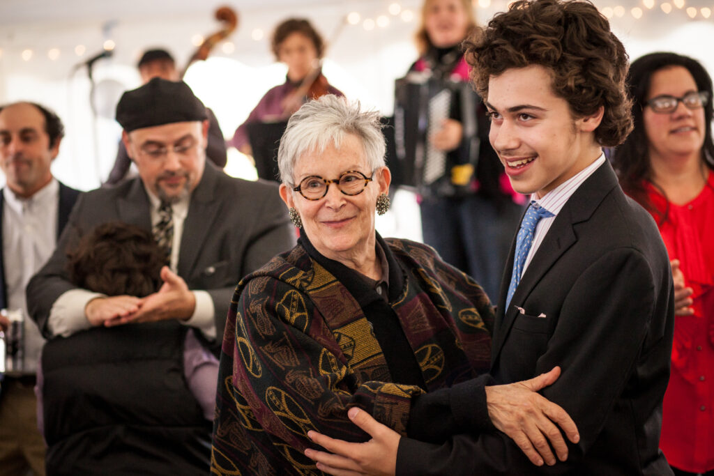 An older woman and a young man in formal attire dance together at a lively indoor event with other people and musicians in the background.