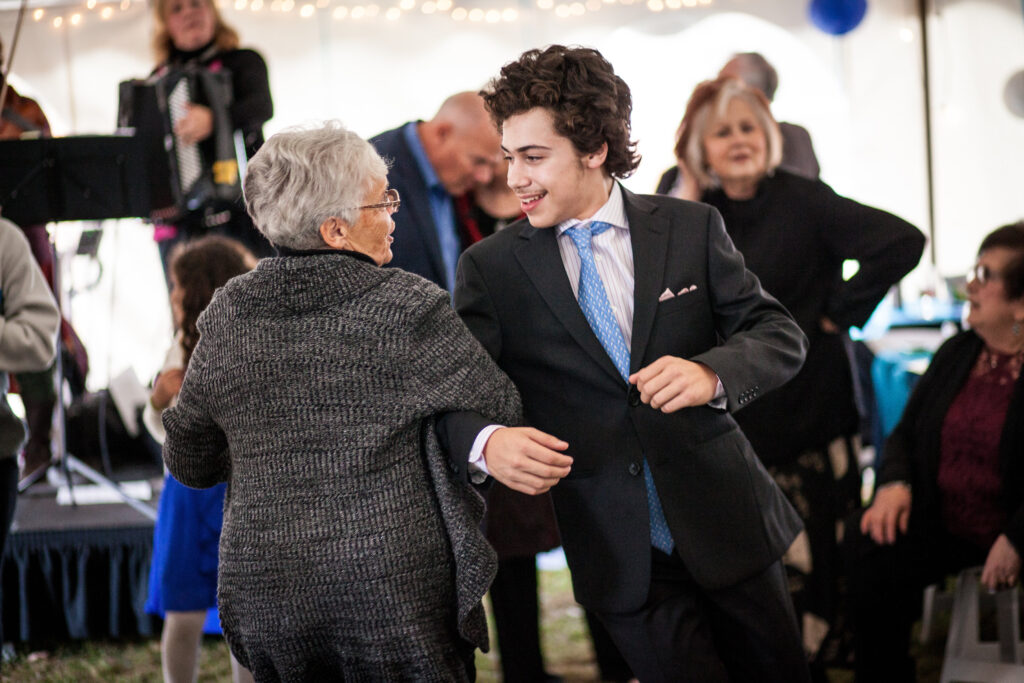 An elderly woman and a young man in a suit dance together at a lively indoor event, with other people and musicians visible in the background.