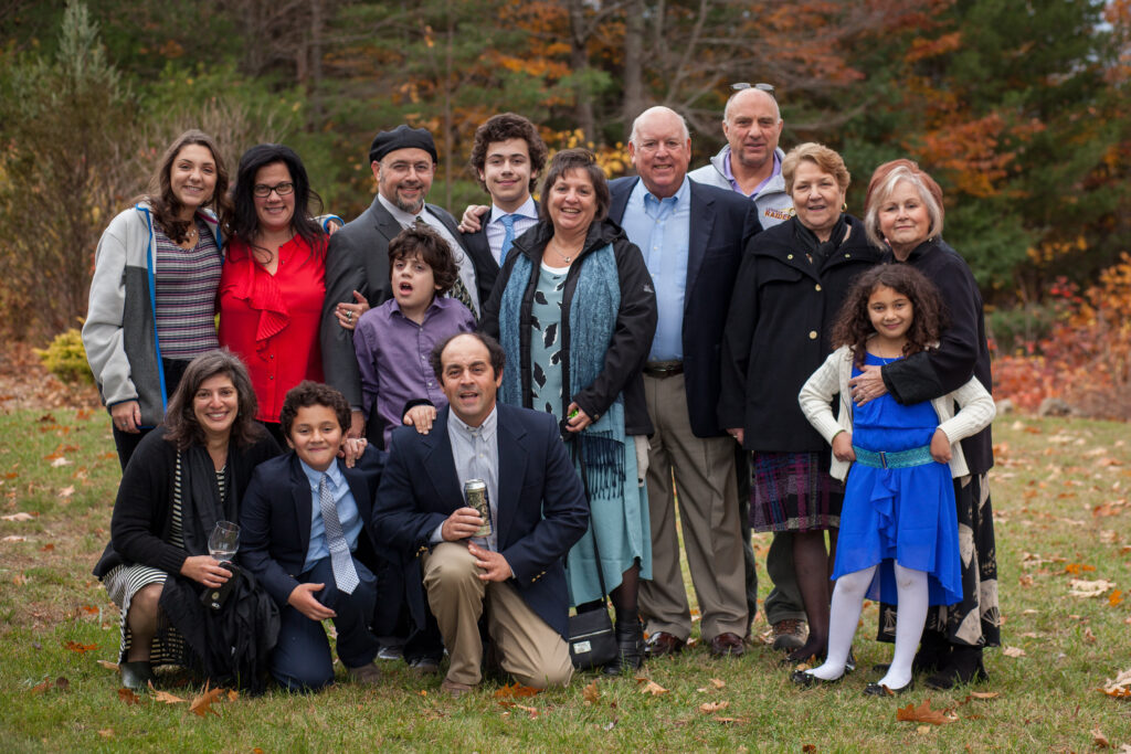 A group of thirteen people, including children and adults, pose together outdoors in autumn, dressed in semi-formal attire and smiling at the camera.