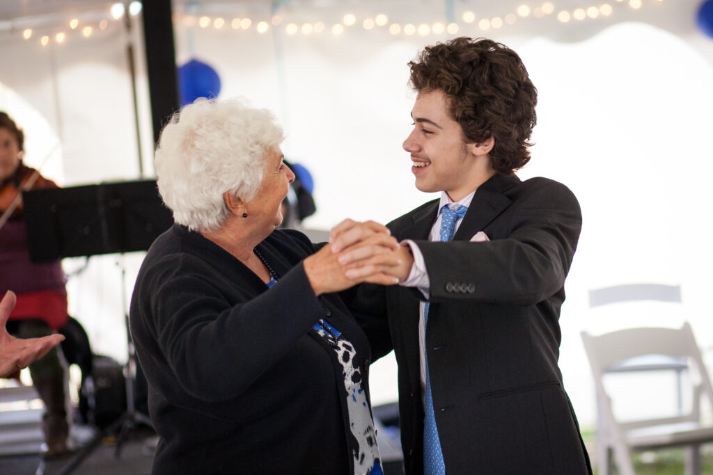 An elderly woman and a young man in formal attire dance together indoors, smiling at each other.