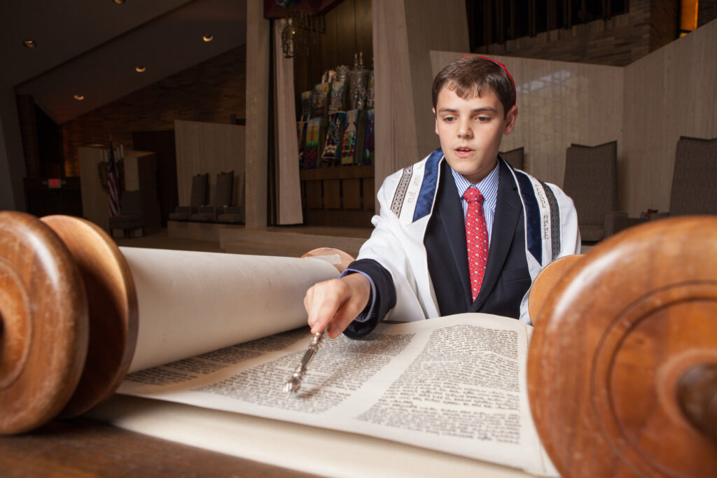 A boy wearing a suit, tallit, and kippah reads from a Torah scroll in a synagogue, using a yad to follow the text.