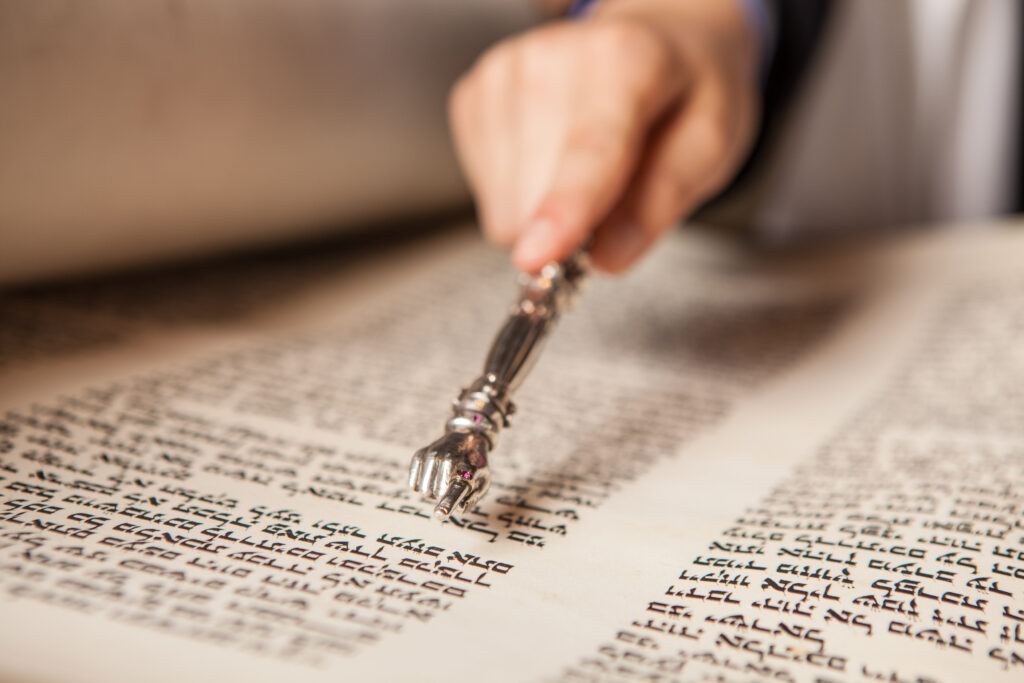 A hand uses a silver pointer to read Hebrew text on a scroll.