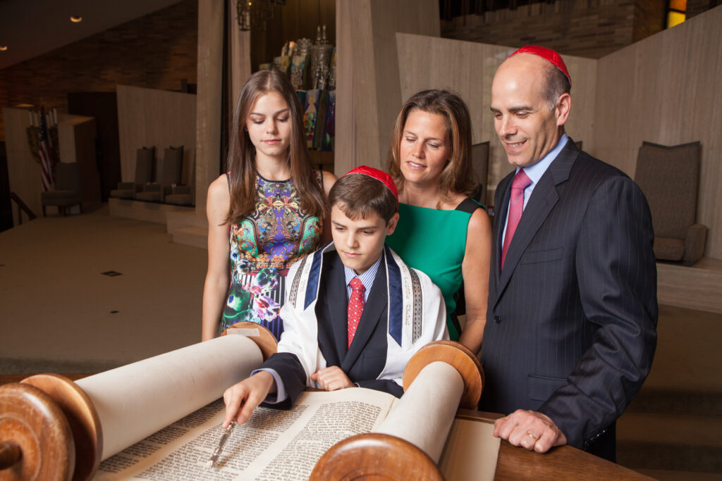 A boy in a tallit reads from a Torah scroll, using a yad, with three family members standing beside him in a synagogue.