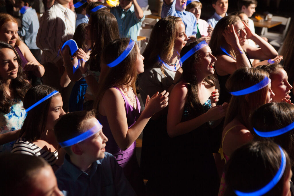 A group of young people wearing blue glow stick headbands stand close together at a lively indoor event, clapping and smiling.