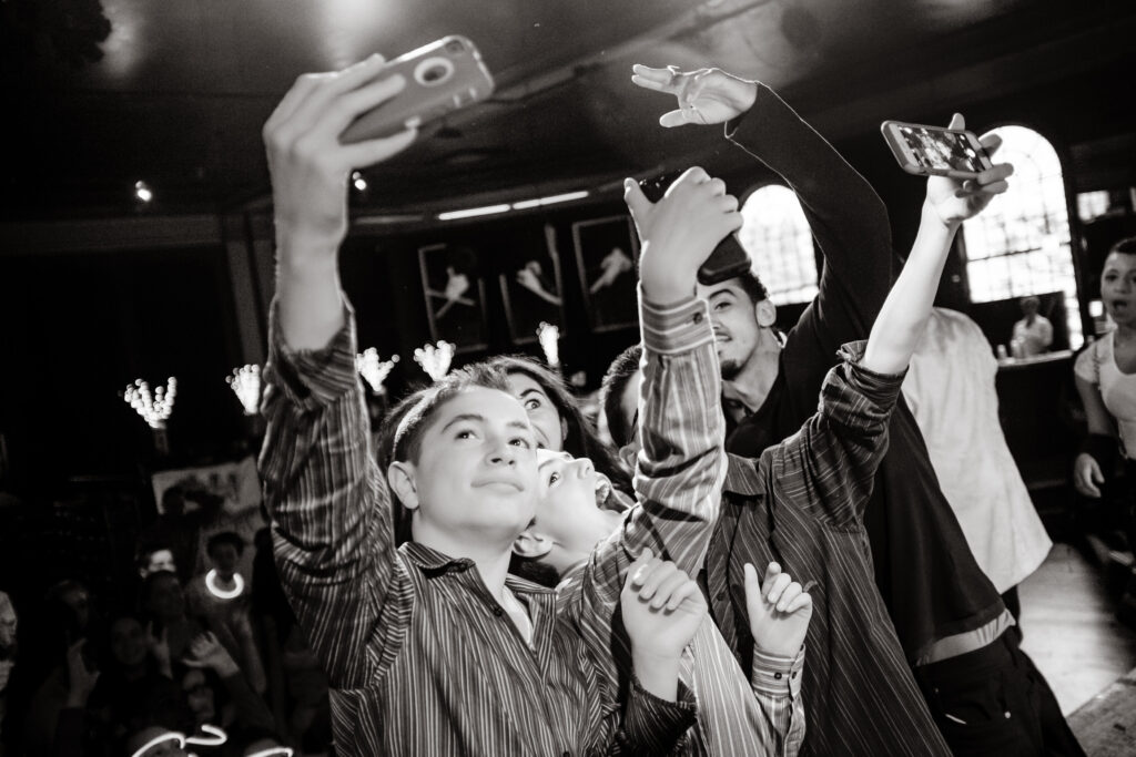 A group of people pose closely together, taking selfies with their smartphones at an indoor event. The scene appears lively and social.