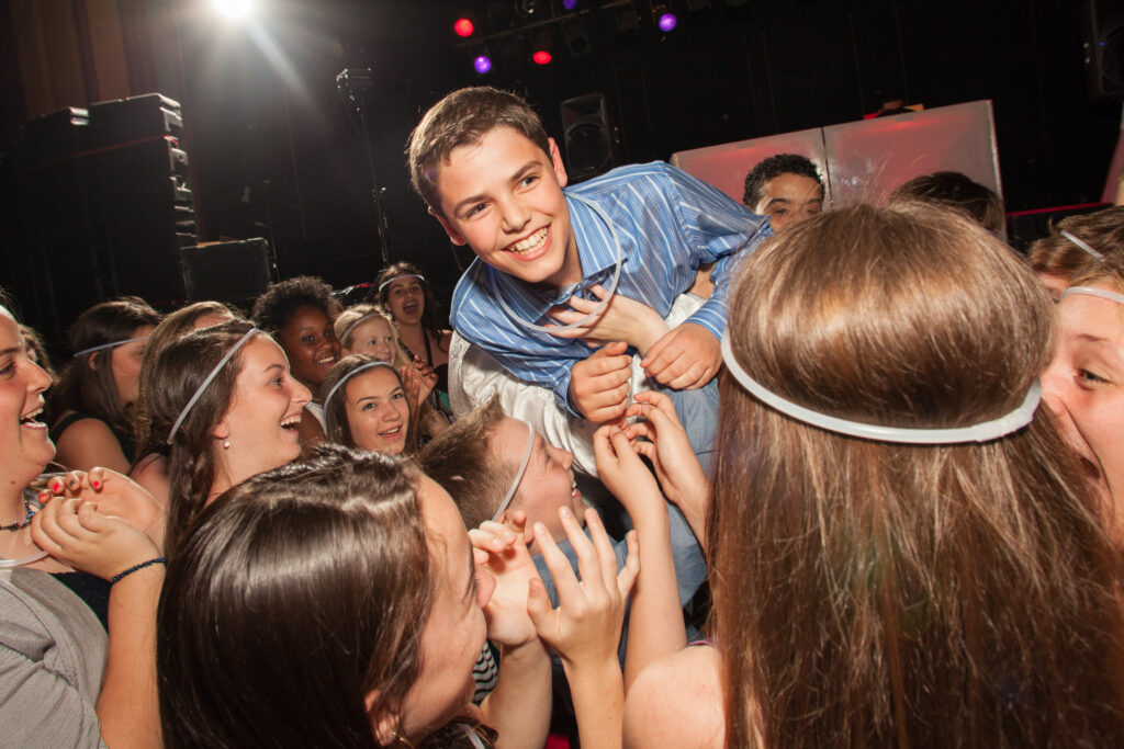 A group of teenagers lifts a smiling boy wearing a striped shirt above their heads in a crowded, lively indoor celebration.