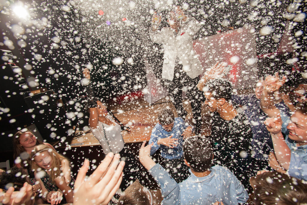 A crowd of people dance under artificial snowflakes at an indoor event, with hands raised and bright lights in the background.