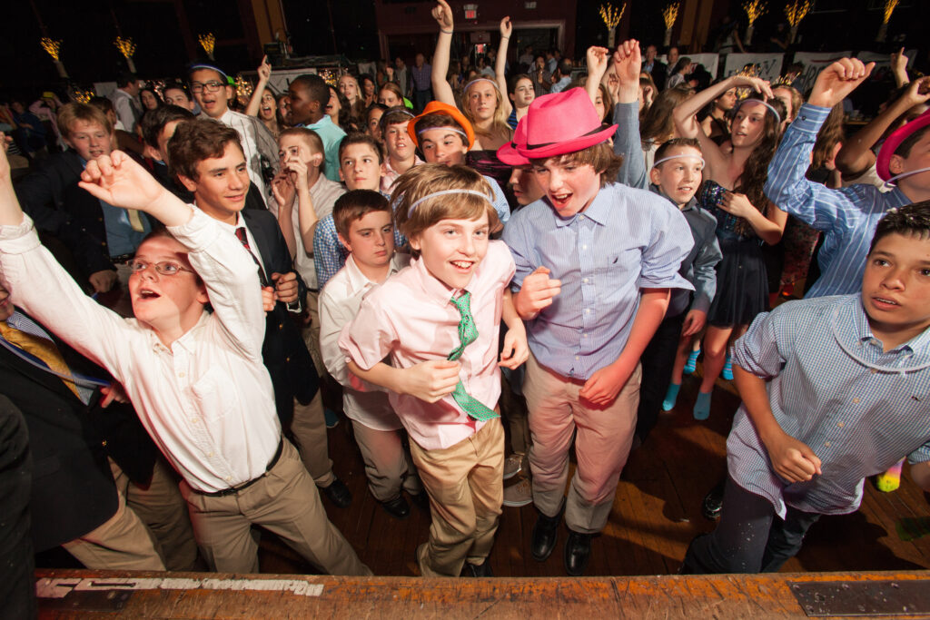 A group of children, mostly boys, dressed in semi-formal clothes, enthusiastically dance and cheer at a lively indoor party or event.