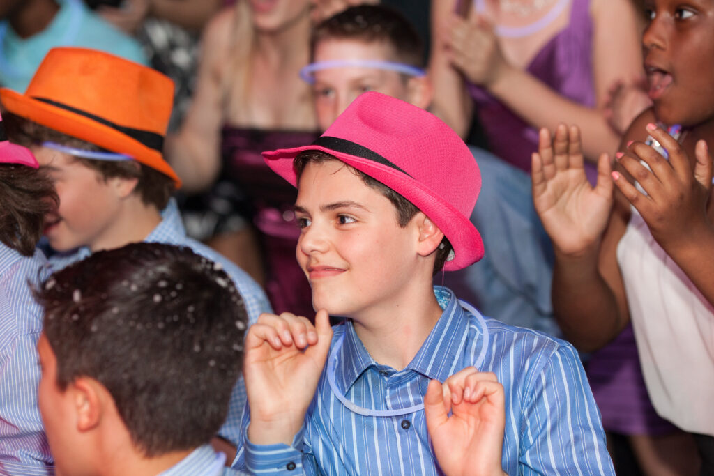 A group of children in colorful hats and glow necklaces clap and smile at a lively indoor event.