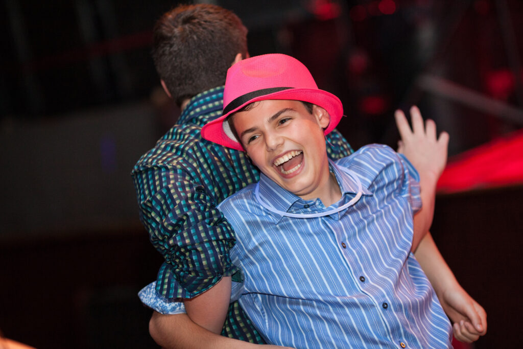 Two boys, one in a pink hat and striped shirt, stand back-to-back and smile widely in a lively indoor setting.