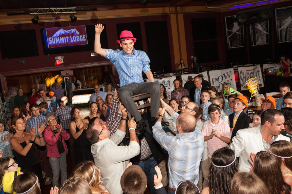 A teenage boy in a pink hat is lifted on a chair by adults at a crowded party, while guests watch and celebrate in a decorated indoor venue.