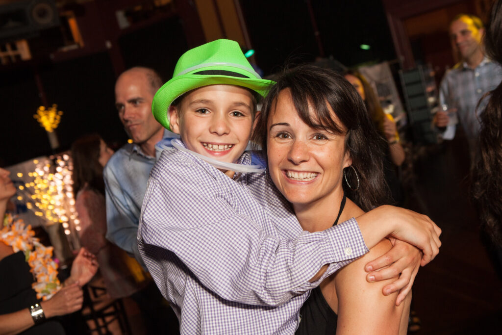 A smiling woman holds a happy child wearing a green hat at a lively indoor event with other people in the background.