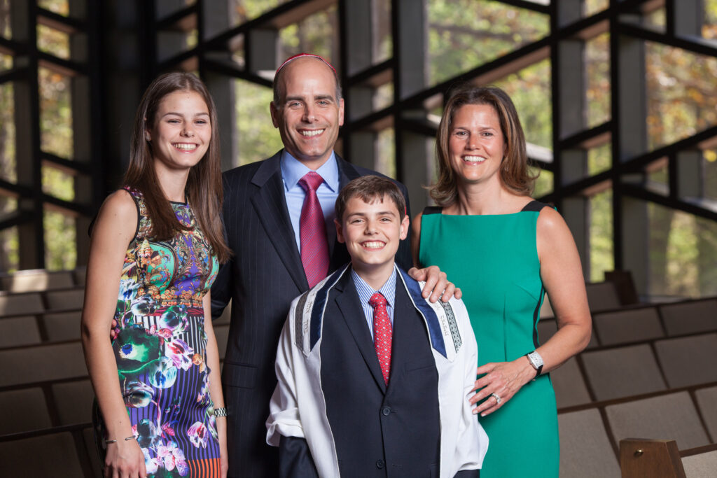Four people, two adults and two children, stand smiling together in a synagogue, with wooden benches and large windows in the background.