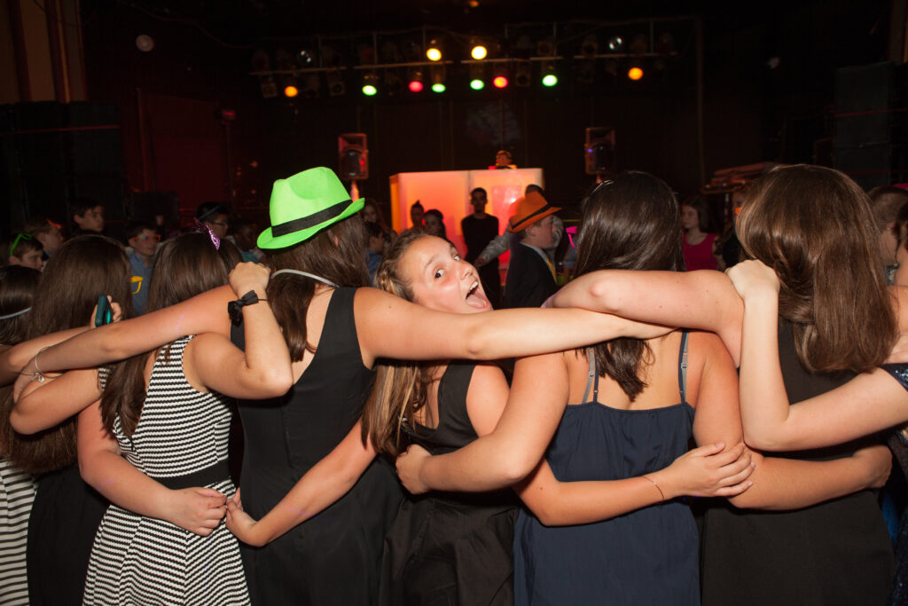 A group of people with arms around each other stand in a row at a party. Some wear hats, and colorful stage lights are visible in the background.