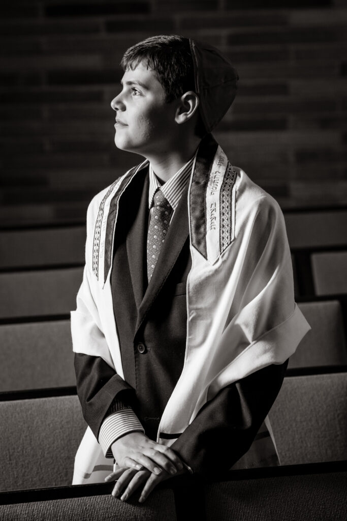 A young man wearing a suit, tie, prayer shawl, and yarmulke stands in a synagogue, looking to the side with hands resting on a pew. Black and white photo.