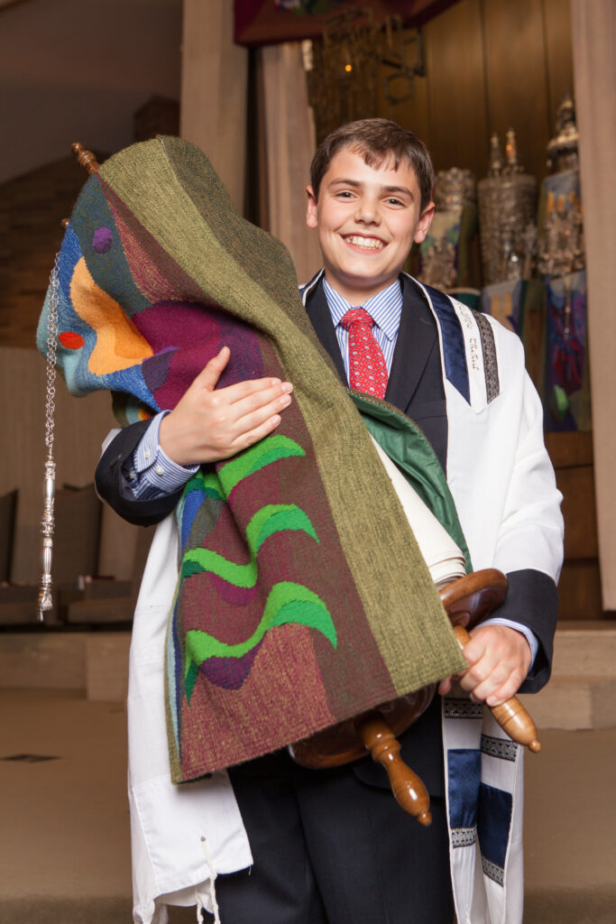 Smiling boy in formal attire and prayer shawl holding a Torah scroll with a colorful cover, standing inside a synagogue.