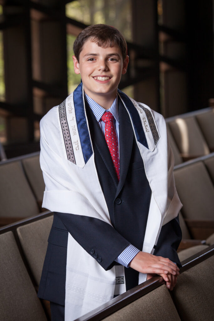 A boy wearing a suit, red tie, and tallit stands smiling in a synagogue, with pews and windows in the background.