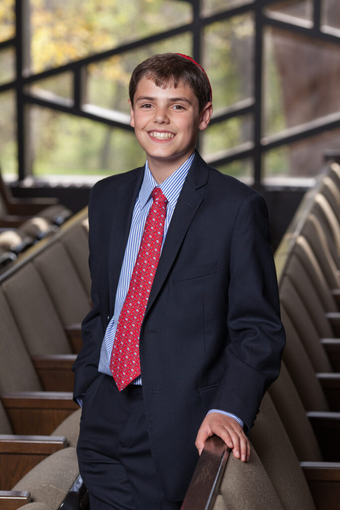 A boy in a suit and red tie stands and smiles in a row of empty seats, with large windows and trees visible in the background.