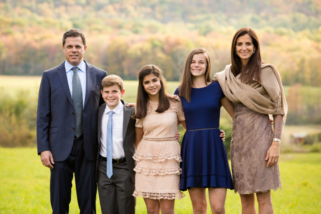 Five people, two adults and three children, stand together outdoors on grass with trees and hills in the background, dressed in formal attire, smiling at the camera.