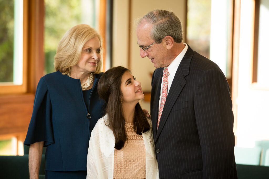 Three people, two adults and one young woman, stand together indoors. The young woman looks up at the man in a suit while the older woman stands beside them, all appearing engaged in conversation.