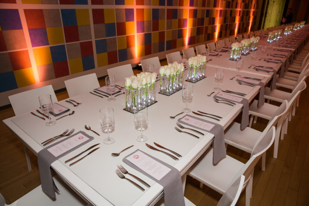 Long banquet tables set with white plates, glassware, silverware, gray napkins, menus, and vases of white roses, against a colorful tiled wall and warm lighting.