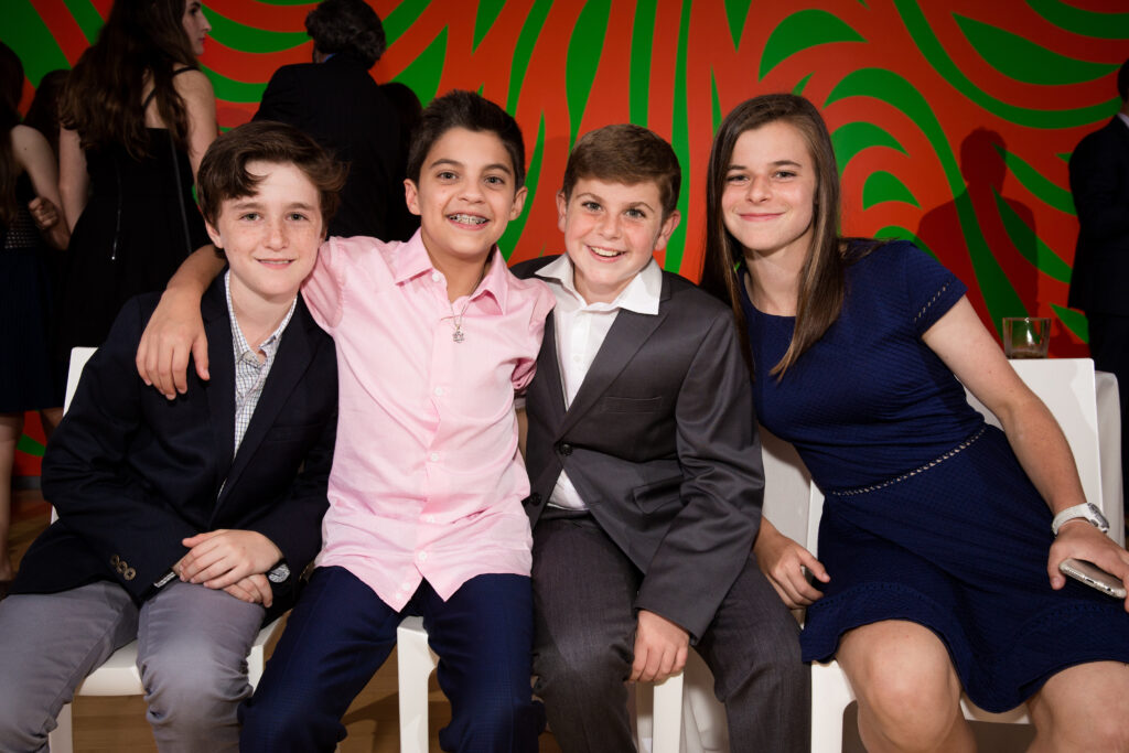 Four kids dressed in semi-formal clothing sit closely together on white chairs, smiling at the camera. The background is a bright red and green abstract pattern.