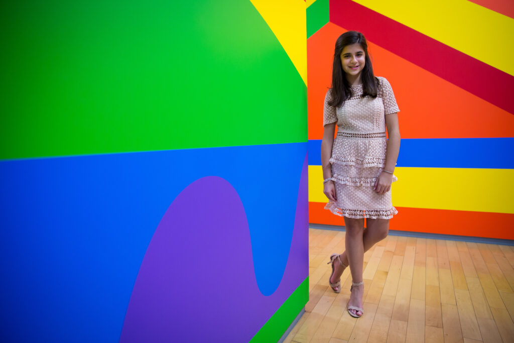 A young woman in a light pink dress stands by a bright, multicolored geometric wall in a gallery with a wooden floor.