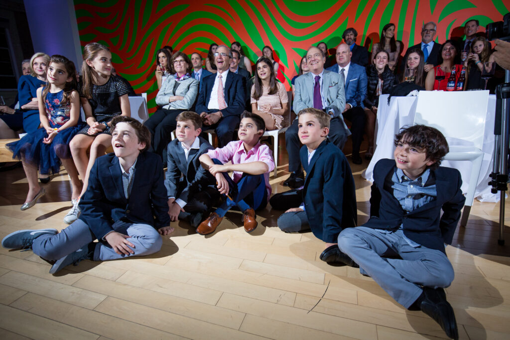 A group of children in formal attire sit on the floor while adults sit behind them, all watching something off-camera in a brightly lit room with a colorful, patterned wall.