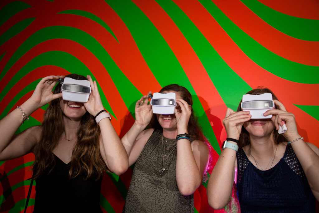 Three people wearing sleeveless tops use virtual reality headsets in front of a red and green wavy striped wall.