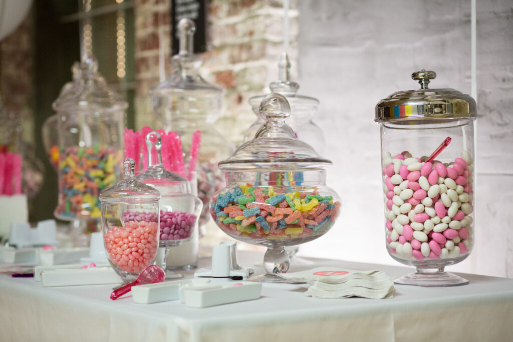 Glass jars filled with assorted colorful candies and gummies are arranged on a white table with scoops and napkins.