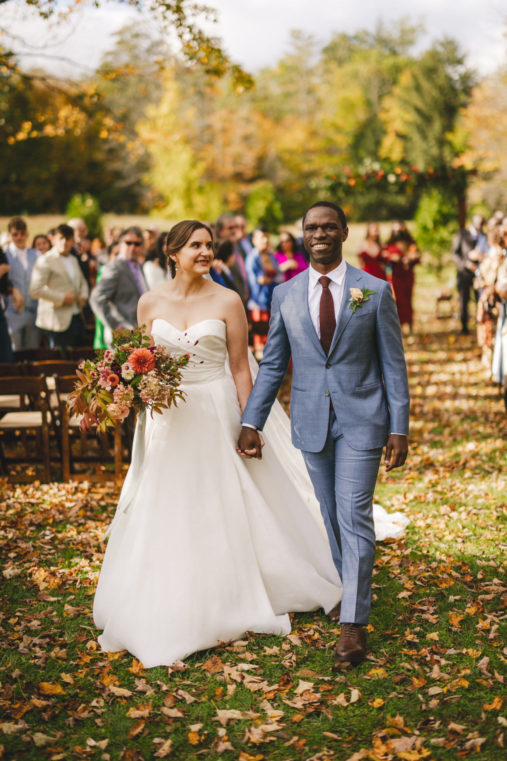 A bride in a white gown and a groom in a light blue suit hold hands and walk down an aisle covered in autumn leaves, captured in Western Massachusetts wedding photography as guests follow behind.
