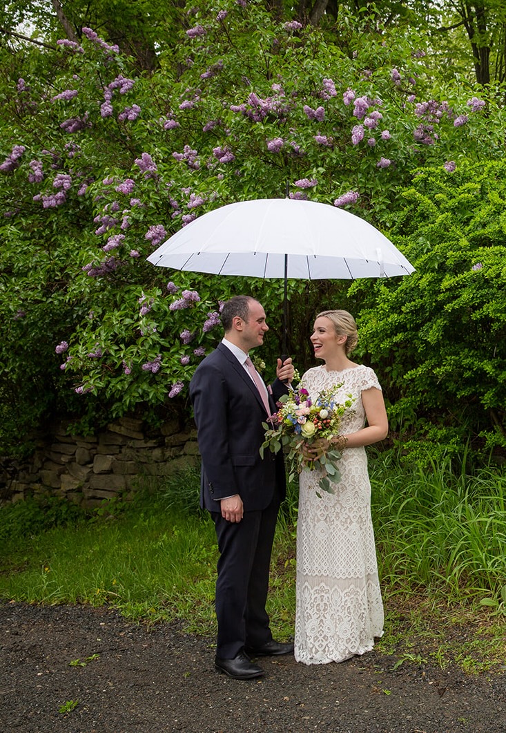 A bride and groom stand under a white umbrella in front of a blooming lilac bush, captured in a style typical of Western Massachusetts wedding photography.