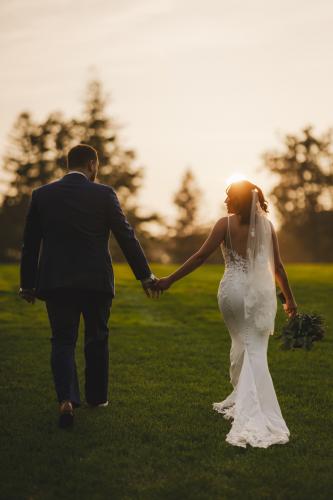 Western MA Wedding Photographer capturing a stunning image of a bride and groom walking through a grassy field at sunset.