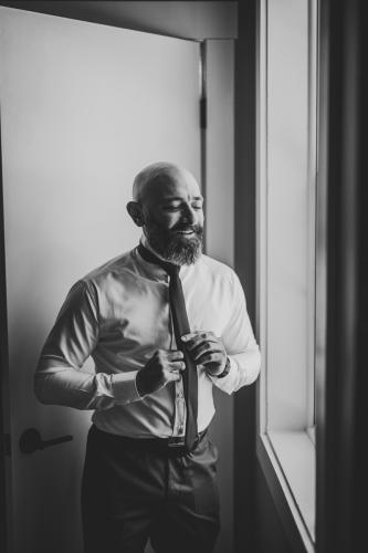 A bearded man getting ready for a Western MA wedding, carefully putting on a tie in front of a window.