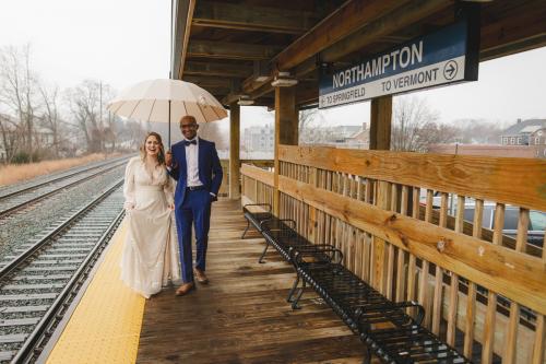 A Western MA Wedding Photographer captures a bride and groom standing on a train platform under an umbrella.